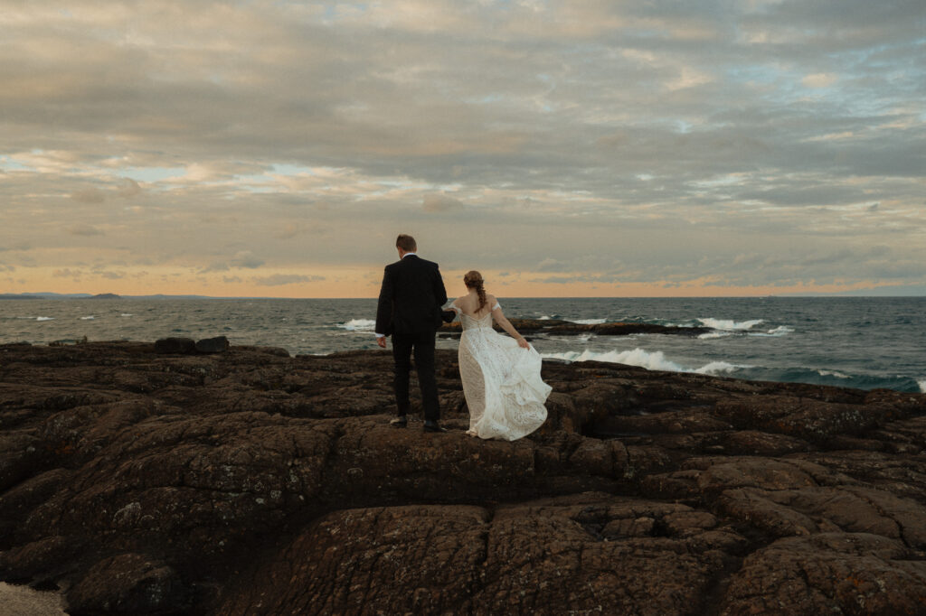 wedding couple walks on black rocks with lake superior waves