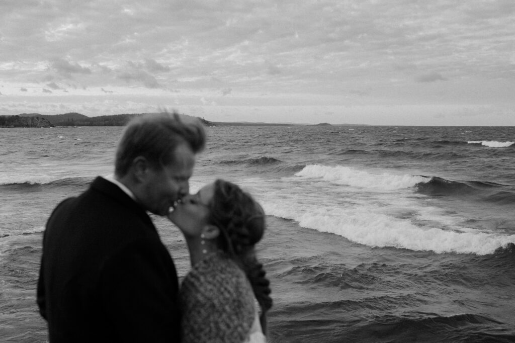 black and white portrait of wedding couple kissing by lake superior waves